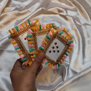 May include: Three square, white cards with gold trim and colorful tassel edges are held in a hand. The tassels are pink, green, orange, and yellow. Small, decorative gems are on the cards.