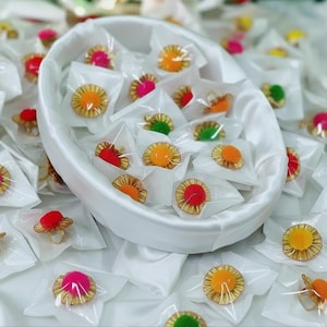 May include: A collection of colorful rings with gold-colored settings and vibrant pom-pom accents, packaged in individual clear plastic bags. The rings are displayed in a white oval-shaped container, surrounded by a white satin fabric.