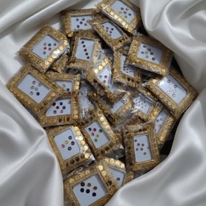 May include: A collection of small, rectangular frames with gold sequin borders and white interiors. Each frame contains colorful, round embellishments. The frames are nestled in a bed of white, silky fabric.