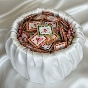 May include: A white satin basket overflowing with small, rectangular cards. Each card features a red grape cluster design, framed with gold and colorful borders. The cards are stacked inside the basket.