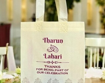 Op de afbeelding: Crèmekleurige draagtas met een handvat. De tas heeft de namen "Tharun" en "Lahari" in een decoratief lettertype, samen met een ringafbeelding. De tekst "BEDANKT DAT U DEEL UITMAAKTE VAN ONZE VIERING" staat ook op de tas.