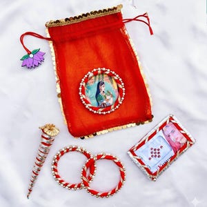 May include: A red drawstring pouch with gold sequins and a picture of a woman. The image also shows red bangles, a henna cone, and a small box with red gems. A purple flower charm is attached to the pouch.