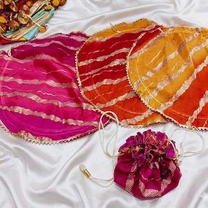 May include: A collection of colorful, round fabric items with gold accents. The items include a drawstring pouch and several circular pieces in shades of pink, red, and orange, with gold stripes and trim. The items are arranged on a white satin surface.