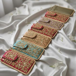 Puede incluir: Colección de carteras de mano rectangulares adornadas en varios colores, incluyendo rojo, dorado y turquesa. Cada cartera presenta intrincados bordados florales dorados y un cierre de solapa a juego. Las carteras están dispuestas sobre una superficie de satén blanco.