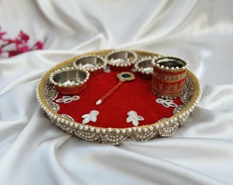 May include: A decorative round tray with a red velvet center, adorned with small silver bowls, a spoon-like object, and white floral accents. The tray's edge is embellished with pearls and gold trim.