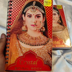 May include: Spiral bound notebook with a woman in traditional Indian bridal attire on the cover. The cover has a red background with gold polka dots and the text "Crystal Designer Spiral Book".