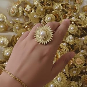 May include: A gold-colored ring with a floral design and a central pearl. The ring is surrounded by other gold-colored rings with similar designs. A gold bracelet is also visible. The jewelry is set against a soft, cream-colored background.