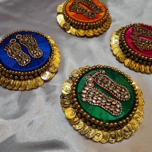 May include: Four decorative, circular ornaments with colorful fabric centers and gold coin edging. Each has a central design of bejeweled footprints. The colors include blue, orange, pink, and green, with gold accents.