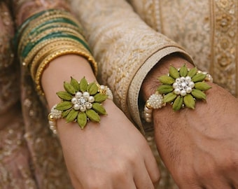 May include: Two flower-shaped bracelets with green petals and pearl accents. The bracelets are worn on wrists, one with green and gold bangles. The bracelets have a central cluster of white pearls and clear crystals. The background shows traditional clothing.