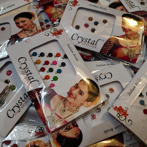 May include: A collection of Crystal Designer Indian Bride bindi packets. Each packet features a variety of colorful, round bindi dots in shades of red, blue, green, and more. The packets also display a woman wearing traditional Indian jewelry.