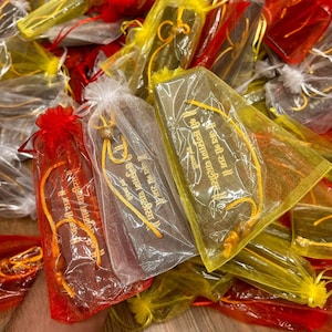 May include: A collection of small, rectangular items wrapped in transparent plastic and tied with colorful ribbons. The items are in red, yellow, and white mesh bags. Gold text is visible on the items, with the words "Hanuman Chalisa" and "Jai Shree Ram".