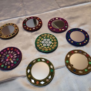 Puede incluir: Colección de ocho pequeños espejos decorativos redondos. Cada espejo está enmarcado con cuentas y adornos coloridos, con una variedad de colores como oro, rojo, verde y morado. Los espejos están dispuestos sobre una superficie blanca.