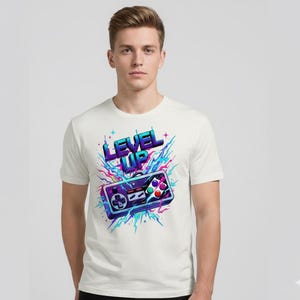 Könnte beinhalten: Weißes T-Shirt mit einem farbenfrohen Grafikdesign eines Retro-Videospiel-Controllers und dem Text "LEVEL UP" in Blau und Lila. Das Design enthält Farbspritzer in Blau, Rosa und Lila.