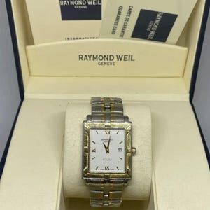 Pode incluir: Um relógio Raymond Weil retangular com moldura dourada e pulseira prateada. O mostrador do relógio é branco com algarismos romanos e ponteiros pretos. O relógio está na caixa original com cartões de garantia.