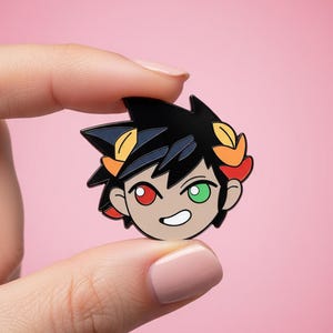 May include: A cartoon pin depicting a character's head with black hair, red and green eyes, and a smiling expression. The pin has orange and yellow accents. It is held between two fingers against a pink background.