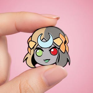May include: A metal enamel pin featuring a stylised character head with split-coloured hair in blonde and dark grey. The character has a light blue crescent moon on the forehead, one green eye, one red eye, and orange accents on the sides of the hair.