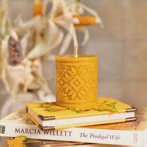 Puede incluir: Una vela amarilla con un patrón geométrico se encuentra sobre una pila de libros. La vela tiene forma cilíndrica y un diseño detallado. Los libros se titulan "MARCIA WILLETT" y "The Prodigal Wife".