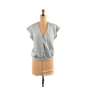 May include: A grey sleeveless cardigan with a V-neck and button fastening. The cardigan has two pockets and a blue embroidered logo on the front.