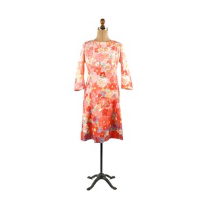 May include: A vintage floral print dress in shades of pink, red, and orange, displayed on a black dress form. The dress features a boat neckline and three-quarter sleeves. The dress is knee-length and has a flared skirt.