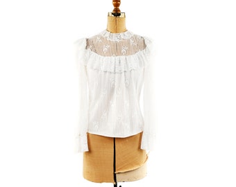 May include: White lace blouse with a high neck and long sleeves. The blouse has a ruffled collar and is on a dress form.
