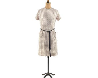 Puede incluir: Vestido vintage en maniquí. El vestido crema tiene un patrón de cuadrícula roja y negra, mangas cortas, cinturón negro y dos bolsillos delanteros. El vestido, a la altura de la rodilla, se exhibe en un maniquí vintage.