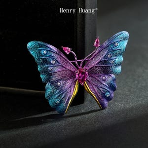May include: A butterfly brooch in shades of blue, purple, and gold, with a central pink gemstone and smaller pink gems on the antennae. The wings are embellished with small crystals. The brooch is branded with "Henry Huang".