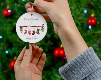 May include: A white ceramic Christmas ornament with the text "Our First Christmas as a Family of Four." The ornament features a brown branch with hanging stockings in red, green, and white. A gold string is attached for hanging.