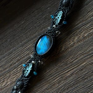 Puede incluir: Pulsera de macramé negro con una piedra ovalada azul de labradorita. La pulsera está adornada con pequeños detalles azules, cuentas plateadas y cuentas de lava negras. El intrincado trabajo de nudos crea una apariencia texturizada y artesanal.
