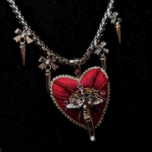 May include: A silver-toned chain necklace featuring a large, red, heart-shaped pendant with embroidered texture. The pendant is adorned with a central silver angel figure. The chain includes multiple silver charms shaped like bows and spikes.