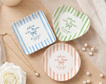 Può includere: Tre piattini in ceramica con bordi dorati: uno quadrato a righe blu con 'future mrs. Moore I said yes! 11.09.26', uno a forma di cuore a righe verdi con 'future mrs. Evans I said yes! 11.08.26' e uno rotondo a righe color pesca con 'future mrs. Reyes I said yes! 12.14.26'.