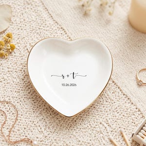 May include: A white heart-shaped ceramic trinket dish with a gold-colored rim. The center of the dish features the black printed text r + t and the date 10.26.2026 in a simple sans-serif font, centered within the smooth, glossy surface.
