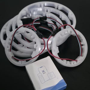 Puede incluir: Una colección de anillos de luz LED blancos y circulares con cables rojos y negros, y una pequeña caja con un diseño azul y blanco. La caja tiene un gráfico de un teléfono y un mando a distancia.