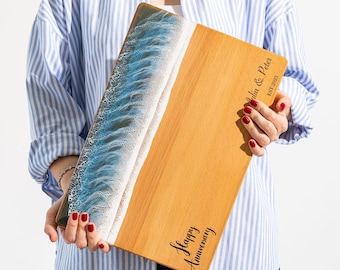 May include: A wooden serving board with a beach-themed design. The board features a blue and white resin wave design on one side and a light brown wood grain on the other. The words "Happy Anniversary" and "Julia & Peter EST 2023" are written on the board.