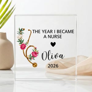 Può includere: Un blocco in acrilico trasparente con una grafica di stetoscopio dorato decorato con fiori rosa e bianchi. Il testo recita THE YEAR I BECAME A NURSE, un cuore nero, il nome Oliva in corsivo e l'anno 2026 stampati in inchiostro nero.