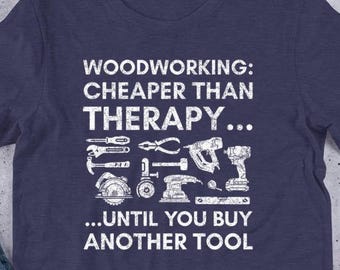 Op de afbeelding: Marineblauw t-shirt met witte tekst en gereedschapsillustraties. De tekst luidt: "Woodworking: Cheaper Than Therapy... Until You Buy Another Tool". Het shirt is gemaakt van zacht materiaal.