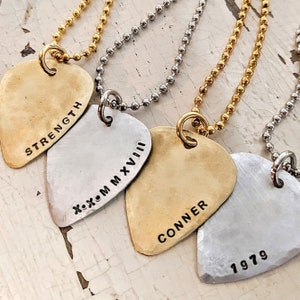 May include: Four silver and gold guitar pick necklaces with different inscriptions. The inscriptions are "STRENGTH", "XOXO MMXVIII", "CONNER", and "1979".