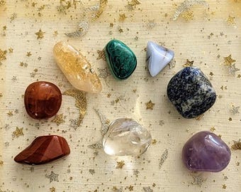 May include: A collection of seven polished gemstones in various colors, including amber, green, blue, and purple. The stones are displayed on a gold-colored, star-patterned fabric pouch.