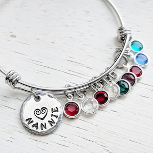 May include: A silver bangle bracelet with a charm that says "Nannie" with a heart and a swirl design. The bracelet has multiple charms with red, green, blue, and clear crystals.