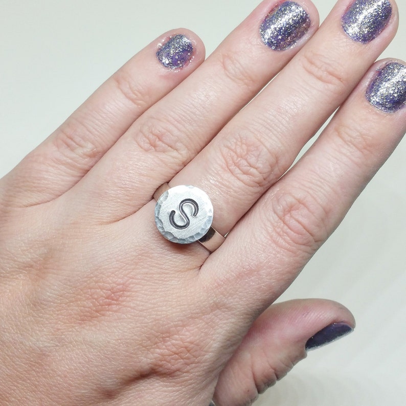 Op de afbeelding: Een zilveren ring met een gehamerde textuur en een cirkelvormige voorkant. De voorkant van de ring heeft de letter "S" in een vetgedrukte, schreefloze lettertype. De ring wordt aan een vinger getoond.