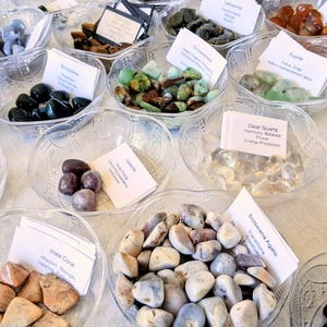 May include: A variety of polished gemstones displayed in clear, decorative bowls. The stones vary in color, including blue, green, brown, and clear. Each bowl has a small white card with text. The arrangement is on a white surface.