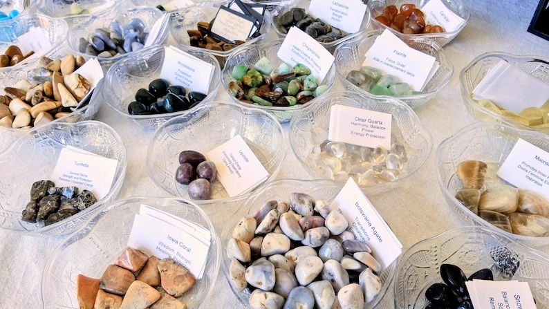 May include: A variety of polished gemstones displayed in clear, decorative bowls. The stones vary in color, including blue, green, brown, and clear. Each bowl has a small white card with text. The arrangement is on a white surface.