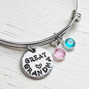 May include: A silver bangle bracelet with a charm that reads "Great Grandma" with a heart in the middle. The bracelet has two small charms with pink and blue crystals.