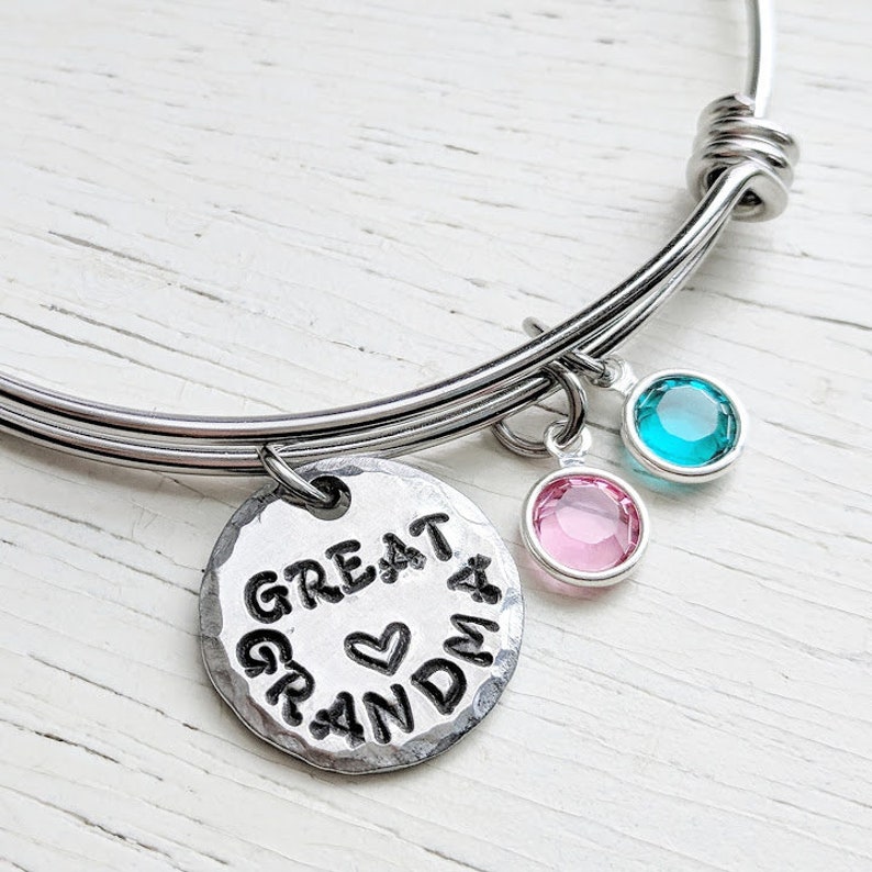 May include: A silver bangle bracelet with a charm that reads "Great Grandma" with a heart in the middle. The bracelet has two small charms with pink and blue crystals.