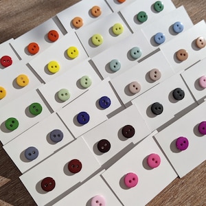 Może przedstawiać: Zestaw kolorowych guzików w różnych rozmiarach i kształtach, ułożonych na białym tle. Guziki są wykonane z różnych materiałów, w tym z tworzywa sztucznego, metalu i drewna.