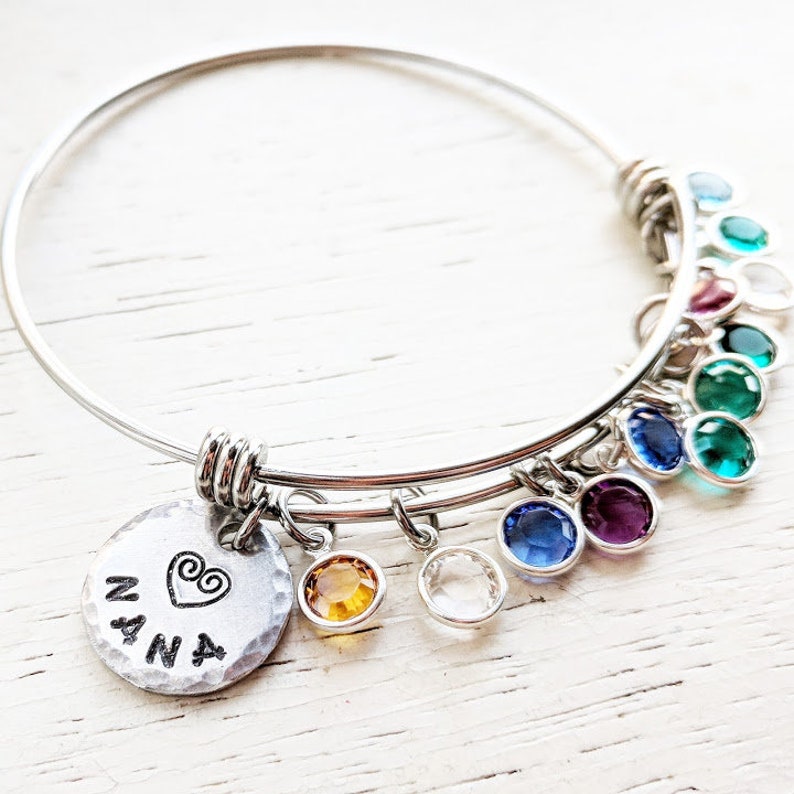 May include: A silver bangle bracelet with a charm that says "NANA" with a heart. The bracelet has multiple colorful crystal charms hanging from it.