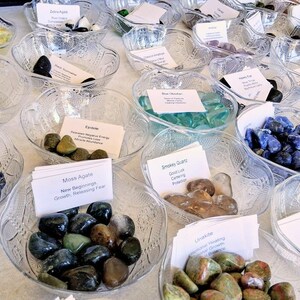 May include: A display of various polished gemstones in clear, decorative glass bowls. Each bowl contains a different type of stone, such as blue lapis lazuli, green moss agate, and clear quartz. Small white cards with handwritten text accompany each bowl.