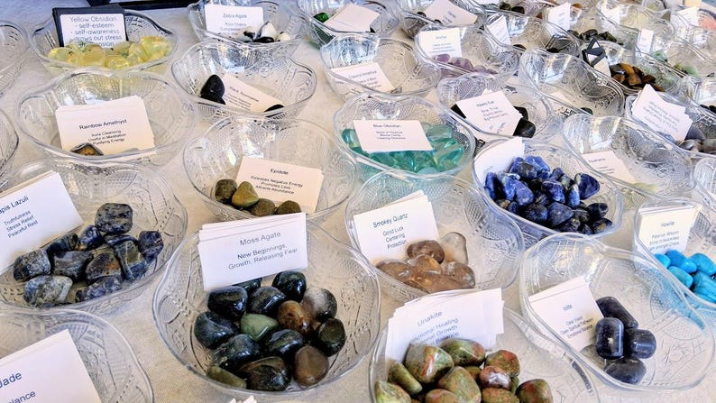 May include: A display of various polished gemstones in clear, decorative glass bowls. Each bowl contains a different type of stone, such as blue lapis lazuli, green moss agate, and clear quartz. Small white cards with handwritten text accompany each bowl.