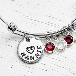 May include: A silver bangle bracelet with a charm that says "Nannie" with a heart design. The bracelet has red and clear crystal charms.
