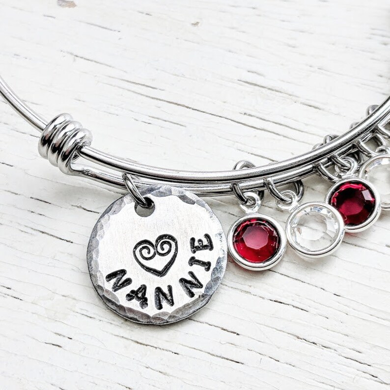 May include: A silver bangle bracelet with a charm that says "Nannie" with a heart design. The bracelet has red and clear crystal charms.