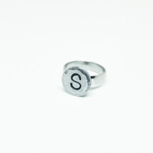 Op de afbeelding: Een zilveren ring met een gehamerde, cirkelvormige voorkant. De voorkant heeft de letter "S" in een vetgedrukte, zwarte letter. De ring staat tegen een witte achtergrond, wat het eenvoudige ontwerp benadrukt.